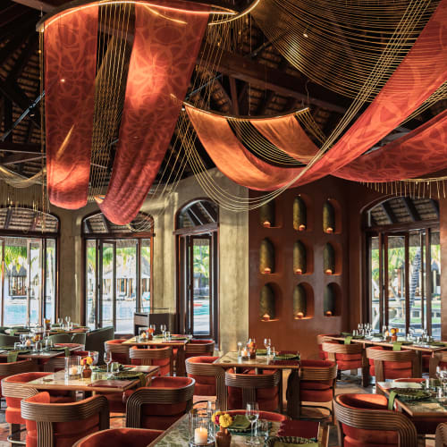 Upscale restaurant interior with curved wooden chairs upholstered in red fabric, marble-topped tables set with glassware and candles, and decorative hanging red fabric with gold thread accents under a high thatched ceiling.