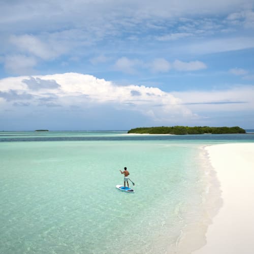 A person paddleboarding on clear turquoise water near a white sandy beach with small green islands in the distance under a partly cloudy sky.