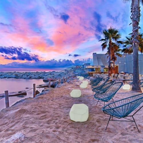 Seaside outdoor lounge area on a sandy beach at sunset with pink and blue clouds, palm trees, modern chairs, and glowing tables.