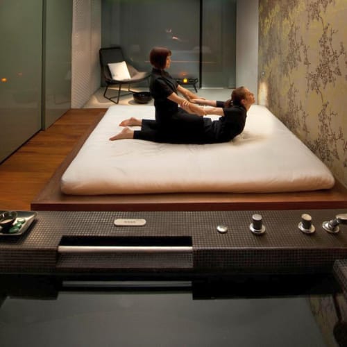 A woman receiving a wellness massage on a mattress in a modern, cozy room with a large window and ambient lighting.