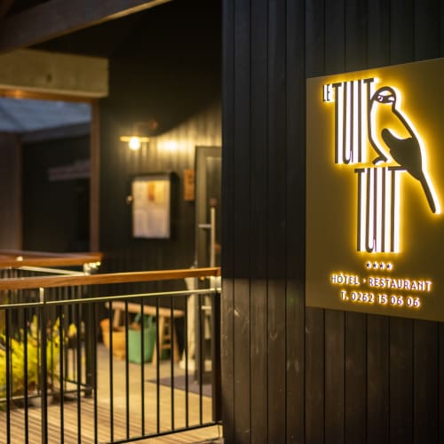 Beleuchtetes Schild von 'Le Tuit Tuit' Hotel und Restaurant an einer dunklen Holzwand bei Nacht, mit Teil eines Decks und Geländern in einer warm beleuchteten Außenumgebung.