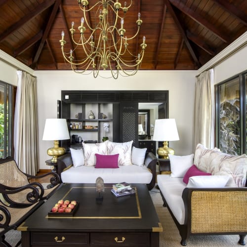 Ein stilvoll eingerichtetes Wohnzimmer mit hohen Decken, einer goldfarbenen-dekorativen Hängeleuchte, großen Glasfenstern, weißen Sofas mit Kissen, einem schwarzen Couchtisch und zwei schwarzen Sessel an den Seiten.