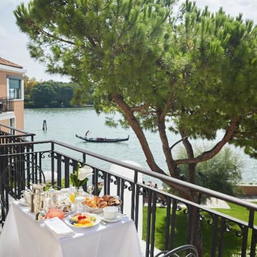 A breakfast setup on a balcony overlooking a river with a gondola, a large tree providing shade, and a picturesque view.