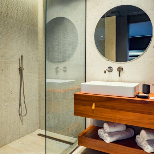 Modernes Badezimmer mit einer glasummantelten Dusche mit rundem Regenduschkopf und einem Holzwaschtisch mit weißem rechteckigem Waschbecken, rundem Spiegel und ordentlich gefalteten weißen Handtüchern auf dem unteren Regal.