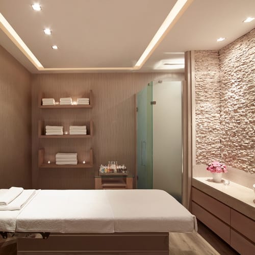Ein moderner Massage- oder Behandlungsraum mit einer Massageliege, Regalen mit Handtüchern, einem Waschbecken und einem dekorativen Spiegel.