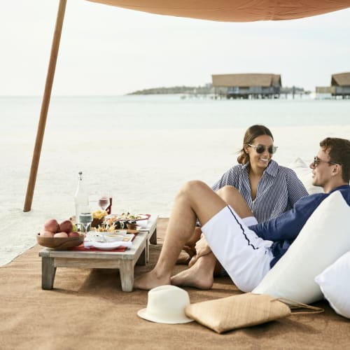 A couple relaxing on the sandbank enjoying a picnic with drinks and snacks under an umbrella.
