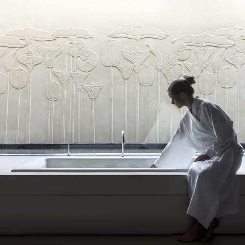 A woman in a white robe sitting on the edge of a large bathroom or spa tub, surrounded by potted plants with white flowers, in a serene and minimalistic setting.