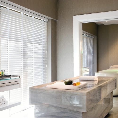 Wellnessraum mit Massagebett, Handtüchern und einem Fenster mit Rollläden neben einer Wand mit ätherischen Ölen.