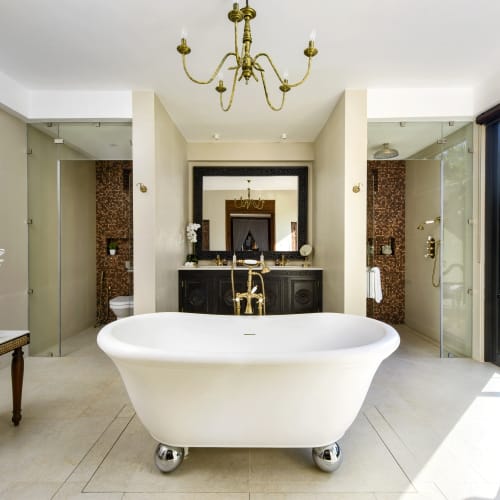 Ein modern ausgestattetes Badezimmer mit freistehender Badewanne in der Mitte, zwei begehbare Duschen auf beiden Seiten, einem großen Fenster, das Tageslicht und eine grüne Landschaft zeigt, und dekorativen Elementen wie einem Spiegel, einer Lampe und Pflanzen.