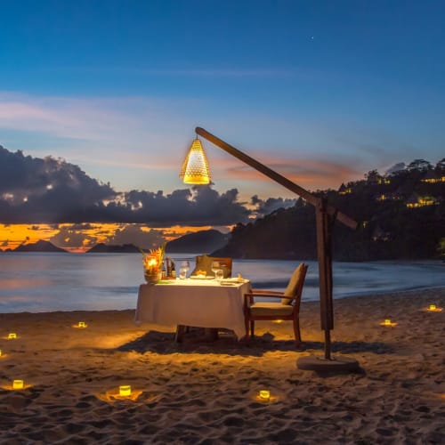 Ein romantisches Abendessen bei Kerzenschein am Strand bei Sonnenuntergang, mit Tisch, Stühlen und Laternen, Blick auf den Ozean.
