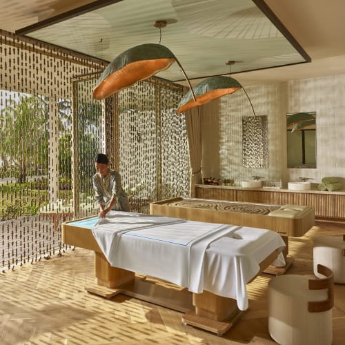 A spa room with two massage tables covered in white sheets, a person preparing one of the tables, and natural light filtering through beaded curtains casting patterned shadows on the floor.