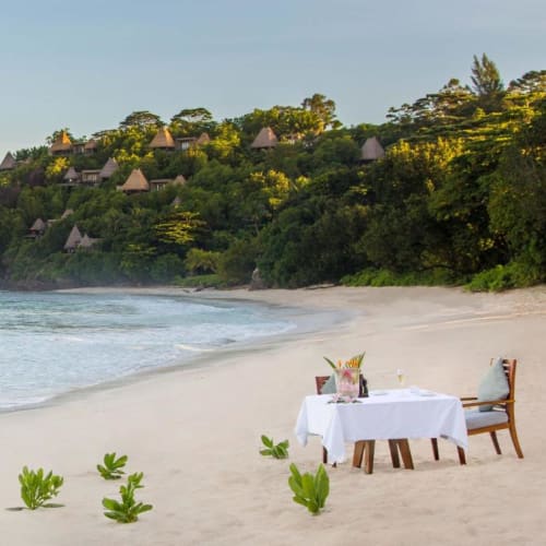Ein privates Abendessen mit Tisch und Stühlen an einem Sandstrand, umgeben von Pflanzen und Blick auf den Ozean.