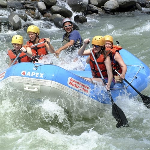 Group of five people rafting on a river with rocks around, all wearing life jackets and helmets, enjoying an adventure.
