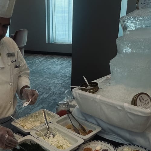 Ein Koch in weißer Uniform und hoher Kochmütze bereitet Teller mit verschiedenen Zutaten neben einer Eisskulptur an einer Kaviarbar in einem Speisesaal vor.