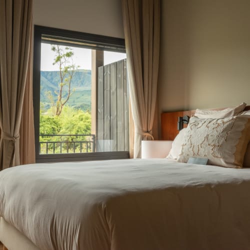 Ein gemütliches Schlafzimmer mit einem ordentlich bezogenen Bett mit beigen Kissen und einem Spitzenkissen, eingerahmt von beigen Vorhängen, mit Blick auf grüne Bäume und einen Berg durch ein großes Fenster.