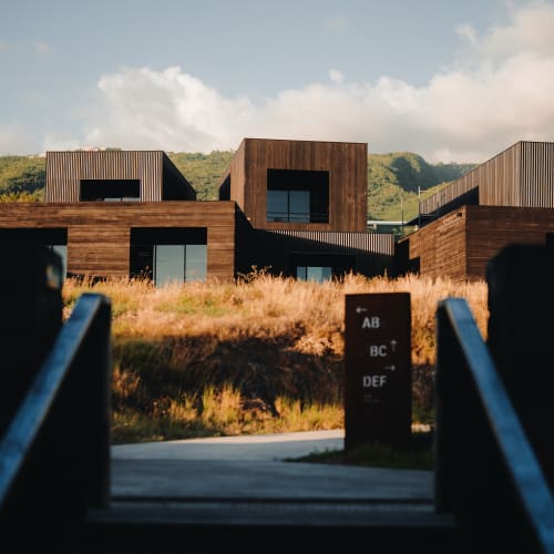 Moderne Holzbauten mit großen Fenstern vor grünen Hügeln unter teils bewölktem Himmel, gesehen von einem Weg, der von dunklen Geländern und einem Wegweiser eingerahmt wird.