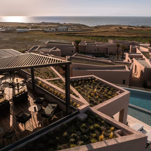 Blick von oben auf ein Dachrestaurant und eine Bar mit moderner Außensitzgelegenheiten unter Pergolen, mit Blick auf einen Pool und weite Felder bis zur fernen Küste bei Sonnenuntergang.