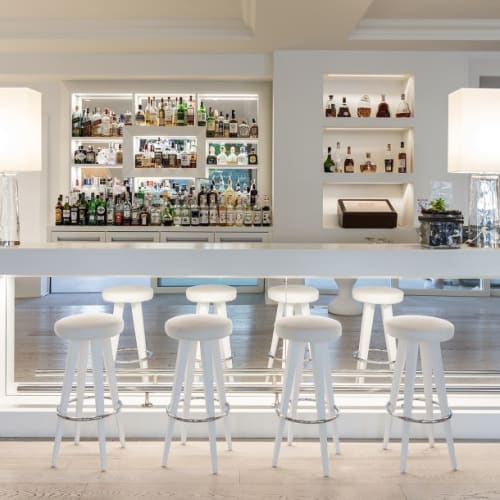 A modern bar with white bar stools and a well-stocked backlit liquor shelf in a luxurious lounge setting.