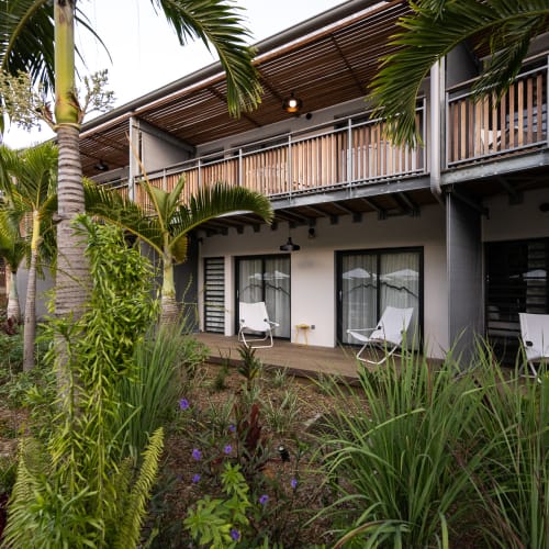 Außenansicht eines modernen zweistöckigen Gebäudes mit Balkonen und großen Fenstern, umgeben von tropischen Pflanzen und Palmen in einer Gartenanlage.