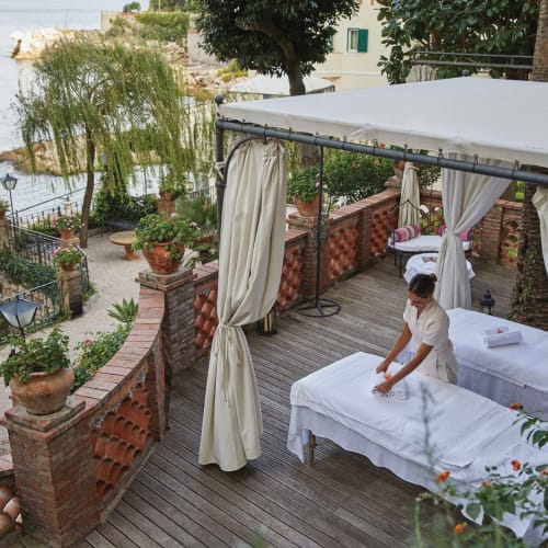 Eine Person erhält eine Massage auf einer umlaufenden Holzterrasse mit Blick auf einen Fluss, umgeben von Topfpflanzen und einem weißen Baldachin.