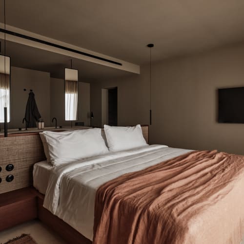 Modernes Schlafzimmer einer Deluxe-Suite mit zwei Schlafzimmern, Doppelbett mit weißer Bettwäsche und beige Decke, wandmontiertem Fernseher und Bettbeleuchtung in einem warmen, neutralfarbenen Raum.