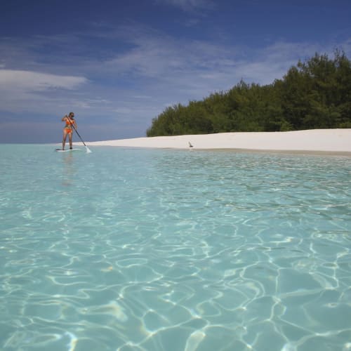 A person stands on a paddleboard in shallow, clear turquoise water near a sandy beach with green trees in the background.