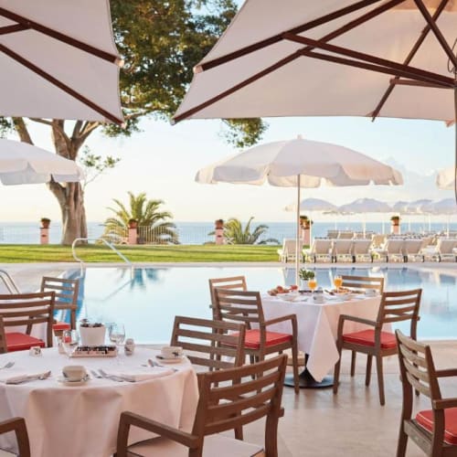 Außerhalb eines Restaurants mit weißen Tischtüchern und roten Kissen um einen Pool mit Sonnenschirmen und Bäumen im Hintergrund.