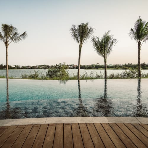 Infinity-Schwimmbad mit Holzterrasse mit Blick auf einen Fluss und mit Palmen gesäumt bei klarem Himmel.