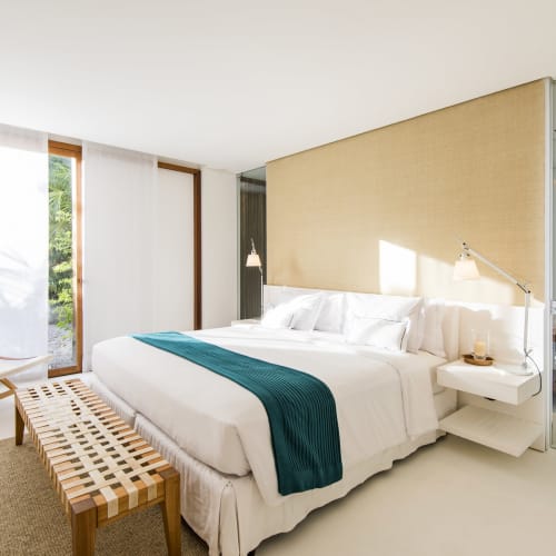 A spacious, bright bedroom with a large bed, a teal throw blanket, and a small sitting area near windows that overlook greenery.