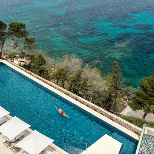 Eine Person schwimmt in einem rechteckigen Außenpool mit Blick auf das Meer, mit weißen Sonnenliegen auf der einen Seite und üppigen grünen Bäumen, die die Gegend umgeben.