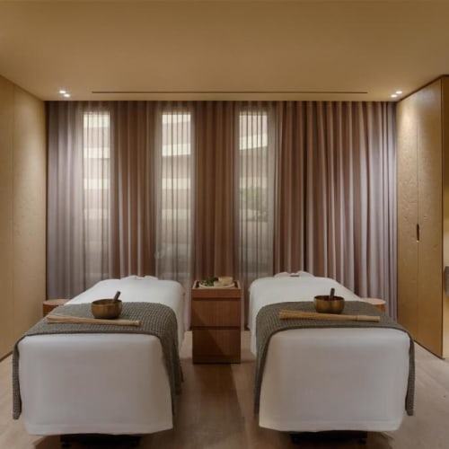 Ein ruhiger Spa-Bereich mit zwei Massageliegen, getrennt durch einen Nachttisch, in einem minimalistischen, gut beleuchteten Raum mit beigen Wänden und transparenten Vorhängen.