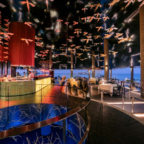 Modernes Innenrestaurant mit einer gebogenen Bar, Tischen mit weißen Tischdecken, vogelartigen Dekorationen an der Decke und großen Säulen mit Bodenbeleuchtung.