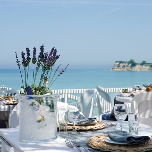 Elegant outdoor dining area with ocean view, decorated with lavender flowers in jars, set for a meal with wine glasses and plates.