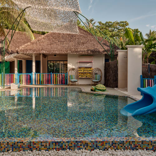 A colorful children's pool area with a blue slide, mosaic tile pool, straw-roofed buildings, and tropical plants in a sunny outdoor setting.