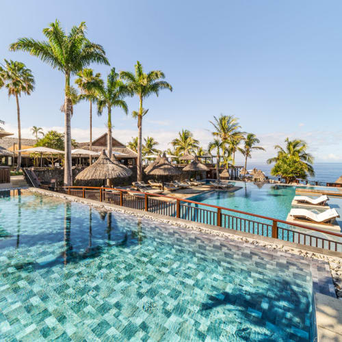 A tropical resort pool area with two adjacent infinity pools, palm trees, thatched umbrellas, and lounge chairs overlooking the ocean under a clear blue sky.