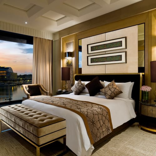 Elegantes Hotelzimmer mit großem Bett, Nachttischen und einer Bank, Blick auf die Stadt und das Wasser bei Sonnenuntergang.
