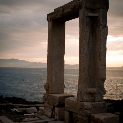 Ancient stone gate ruins at sunset with sea and distant mountains in the background on Naxos island.