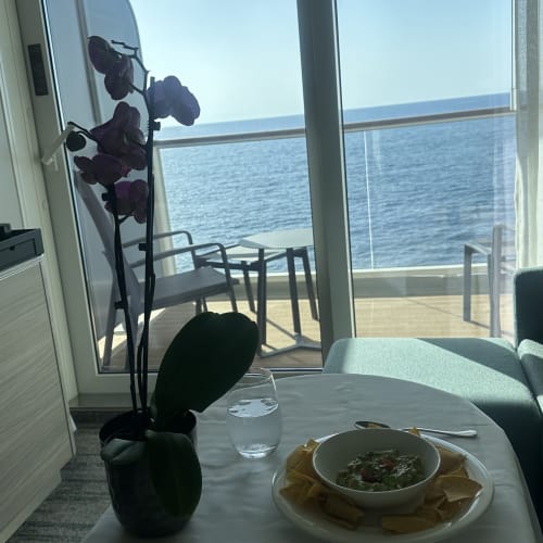 Ein Tisch mit einer Schale Guacamole und Tortillachips, einem Glas Wasser und einer Orchidee in einem Topf in einer Suite mit Blick auf einen Balkon mit Stühlen und Meer im Hintergrund.