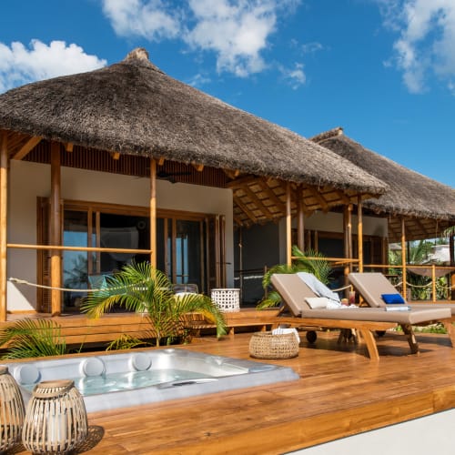 Luxussuite mit Meeresblick, Holzterrasse, Liegen, Whirlpool und tropischen Palmen unter blauem Himmel.