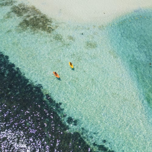 Two people kayaking in the clear waters near a sandy beach, with a dark reef visible in the water.