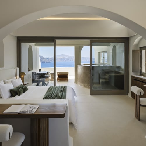 Spacious, modern hotel suite with white walls, a large bed with green cushions and blanket, a wooden desk, a sliding glass door leading to a terrace with outdoor seating, and a view of the sea and mountains.