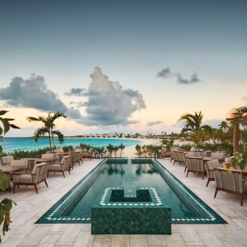 Luxury hotel terrace with a swimming pool overlooking the beach and ocean at sunset, surrounded by lounge chairs and tropical plants.