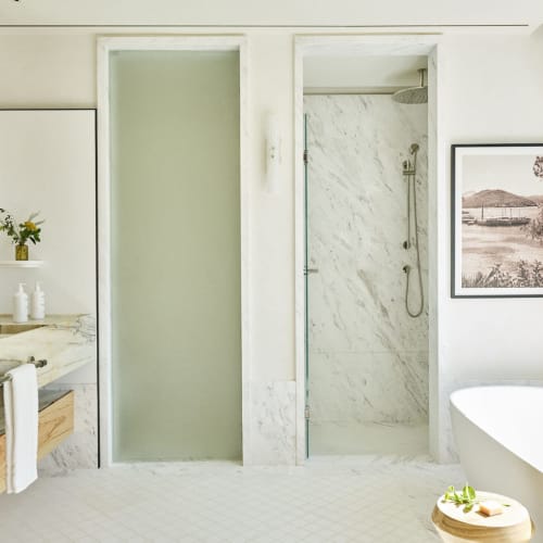 Bright, modern bathroom with marble countertops, a large mirror, a walk-in shower with glass door, a freestanding bathtub next to a window, and wooden accents.