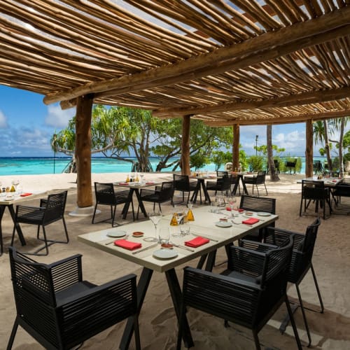 Strandnahes Restaurant mit schwarzen Stühlen und Tischen unter einem Holzdach, mit Blick auf den Strand und den Ozean.