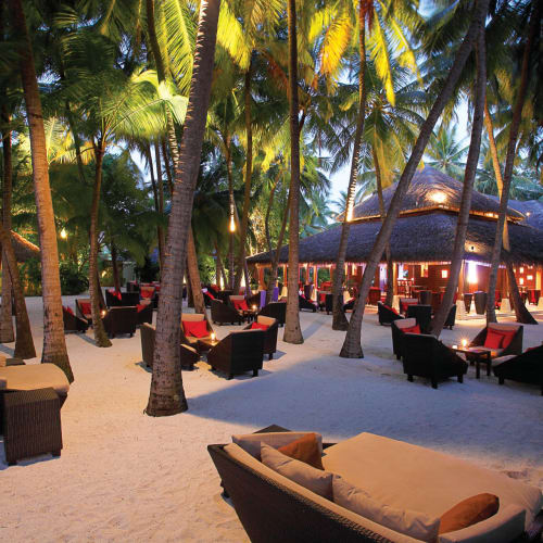 Ein Außen-Loungebereich an einem Strand mit Palmen und beleuchteten Strohdächern, mit Liegestühlen und Sofas.