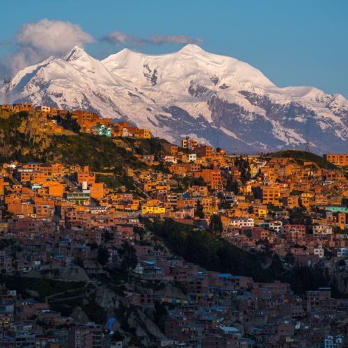 Bunte Häuser in La Paz, die an einen Berghang gebaut sind, mit dem schneebedeckten Illimani-Gipfel im Hintergrund bei Sonnenuntergang.