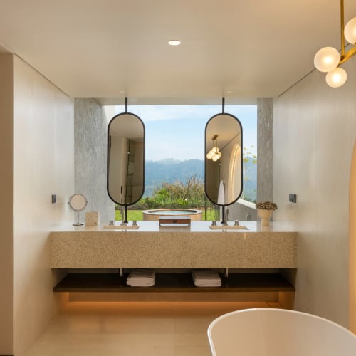 Modernes Badezimmer mit Doppelwaschtisch und zwei ovalen Spiegeln, Blick auf Grünfläche und Berge durch ein großes Fenster und einer freistehenden weißen Badewanne im Vordergrund.