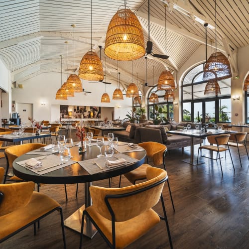 Geräumiges, modernes Restaurantinterieur mit runden und rechteckigen Tischen, gedeckt mit Glasgeschirr und Servietten, ausgestattet mit senffarbenen Stühlen, Korblampen, großen bogenförmigen Fenstern und Holzboden.