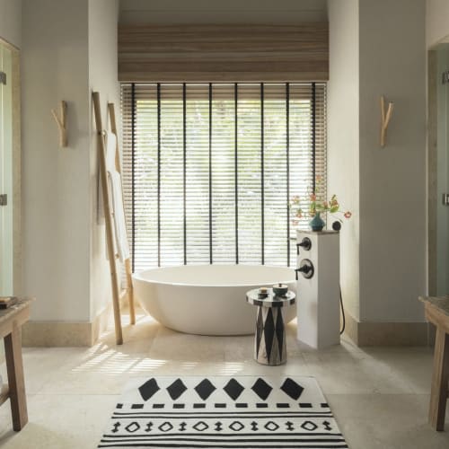 Modernes Badezimmer mit zwei hölzernen Waschbecken, einer runden weißen Badewanne, einem schwarz-weiß gemusterten Teppich und großen Jalousien, die natürliches Licht hereinlassen.
