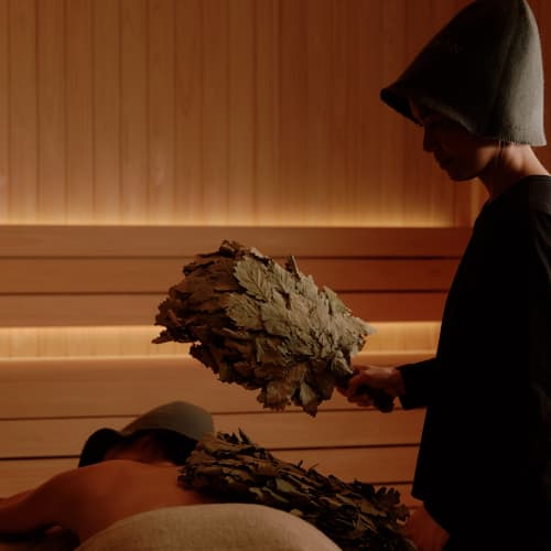 A person in a spa or sauna room, receiving a treatment while another person stands nearby holding two leaf fans.